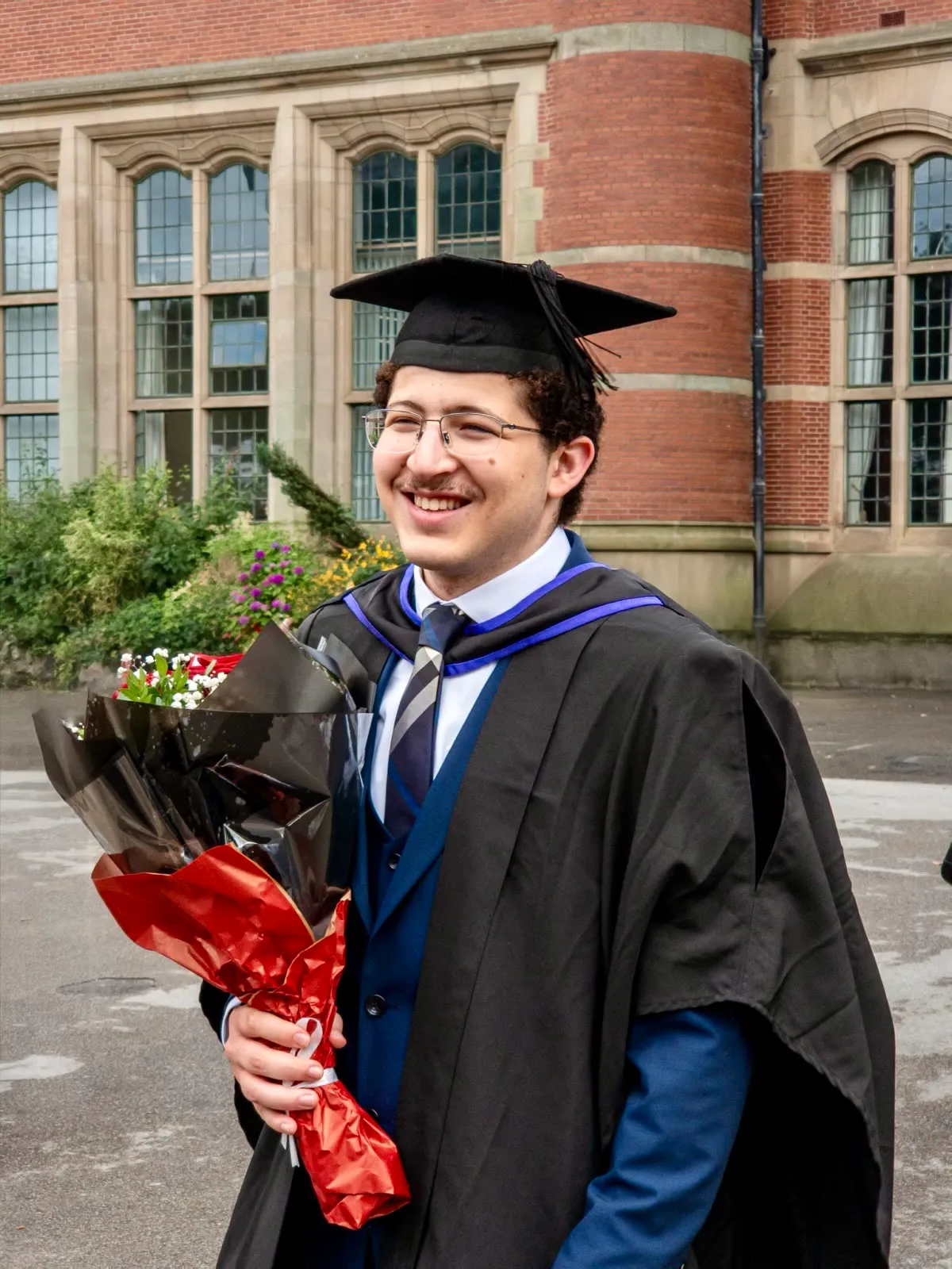 Smiling in my gown, holding flowers, outside one of the university's red-brick buildings